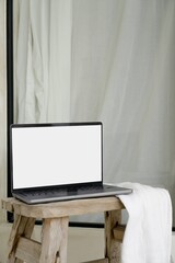 Laptop MacBook Pro computer mockup with white screen on rustic, vintage wooden light brown stool draped with white linen throw in modern, minimal, white Villa in Bali. Bohemian (Boho) and Scandinavian