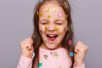 Extremely happy joyful little girl covered colorful kid's stickers standing isolated over gray...