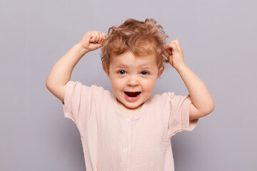 Amazed excited blonde little caucasian infant girl posing isolated over gray background raised arms...