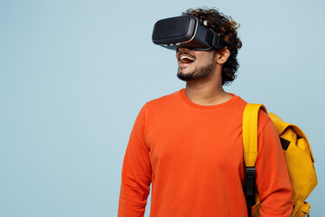 Young fun teen Indian boy student wears casual clothes backpack bag watching in vr headset pc gadget play game isolated on plain pastel light blue background. High school university college concept.