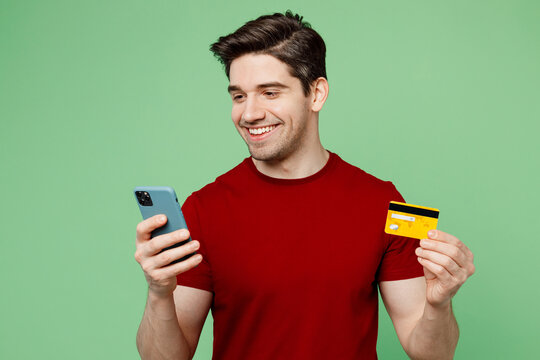 Young man he wearing red t-shirt casual clothes using mobile cell phone hold credit bank card doing online shopping order delivery booking tour isolated on plain pastel light green background studio.