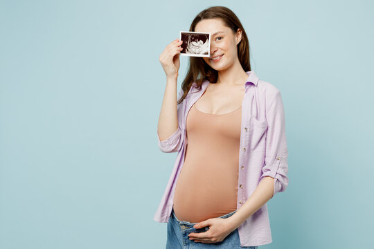 Young pregnant future mom woman with belly tummy wearing casual clothes hold cover eye with ultrasound image pregnant baby photo isolated on plain pastel blue background. Maternity pregnancy concept.