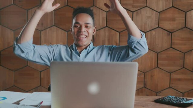Young successful African American man happily waves hand and leans back in chair after completing complex project and fulfilling KPI plan sits in office at table with laptop. Business lifestyle