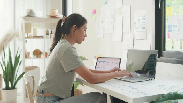 Young Asia girl IT development programmer typing on keyboard coding programming fixing data code on laptop computer screen follow code plan in tablet and paperwork on table workroom at house office.