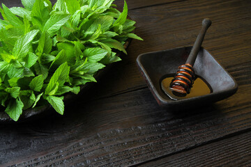 Natural, liquid, yellow, honey in a black, wooden, square plate with a honey dipper, next to it is an oval black wooden plate with fresh mint. And everything lies on a black old wooden table.