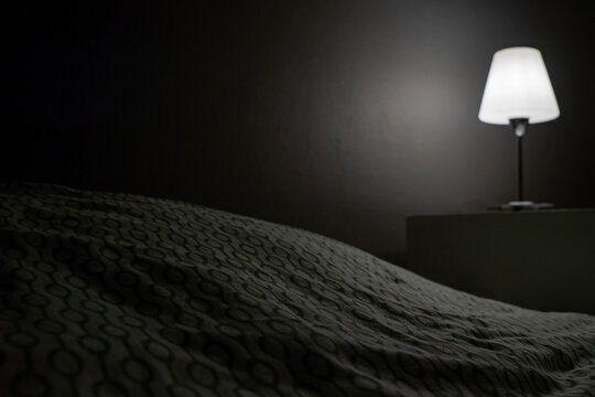 A bedside table and a burning lamp stands on it near the bed in a dark room. Hotel number. Place for text.