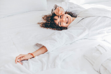 Beautiful cheerful woman in a white bathrobe lying on the bed in the morning. - obrazy, fototapety, plakaty