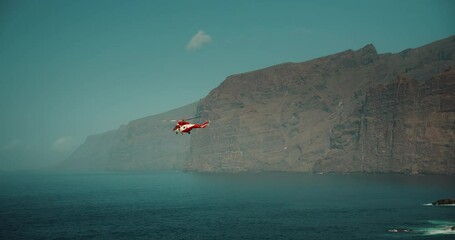 TENERIFE, CANARY ISLANDS, SPAIN, 2023 April: Red medical rescue helicopter flies over a stormy ocean near rocky Los Gigantes cliffs. Generative AI