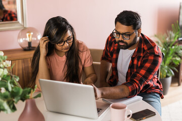 Young millennial indian husband and wife doing domestic paperwork, accounting job and reviewing paper bills, receipts at laptop computer, using online calculator and paying mortgage rent fees on