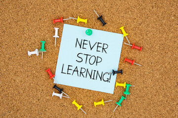 Never stop learning text on light blue post-it paper pinned on bulletin cork board surrounding by...