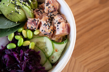 Close-up view of tuna (aku) poke bowl with black rice, fresh cucumbers, red cabbage, avocado, spinach leaves, radish and edamame beans on wooden table in restaurant. Soft focus. Hawaiian food theme.