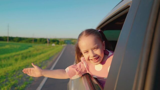 Little girl rejoices in family train by car. Child, stretching his hand out of car window, laughs. Girl child looks out of car window. Happy family travels by car. Childrens emotions. Family vacation