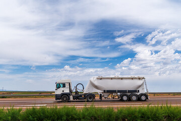 Tanker truck for the transport of bulk cement circulating on a highway under a sky with clouds.