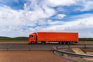 Orange tarpaulin semi-trailer truck driving on a highway under a cloudy sky.