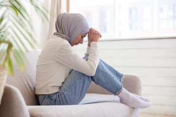 A young Muslim woman in a hijab sits cross-legged on the sofa, resting her head thoughtfully on her...
