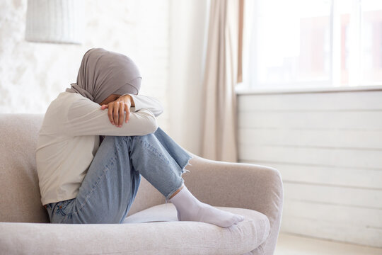 Young Muslim Female Student In Hijab Sitting On Sofa At Home, Resting Head On Knees And Crying. Problems With Studies, Parents, Boyfriend.