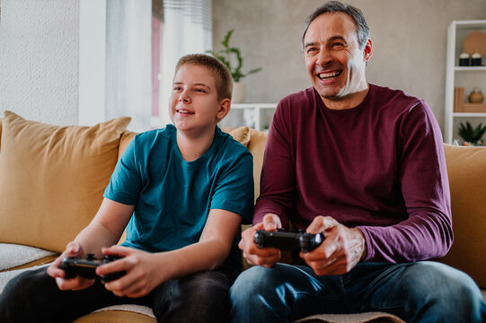 Smiling young boy and his father laughing and playing video games at home.