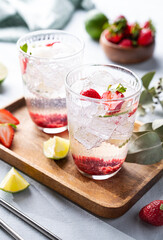 Fresh strawberry mojito in a glass with berries, lime, mint and ice in a wooden plate  on a light background. The concept of a summer refreshing drink for a party