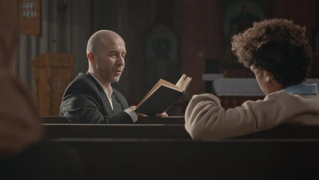 Chest up of middle aged Caucasian bald headed priest with Bible in hands speaking to Biracial teenage boy sitting together on wooden bench in Catholic Church
