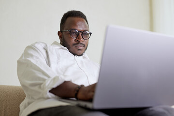 Handsome Black African American Man Working on Laptop Computer while Sitting on a Sofa in Cozy Living Room. Freelancer Working From Home. Browsing Internet, Using Social Networks, Having Fun in Flat.