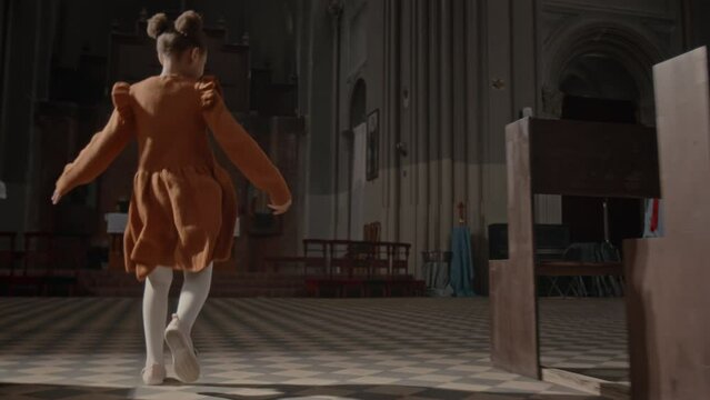 Back view slowmo of little girl in pretty dress and white tights running along central walkway in Catholic Church with delight