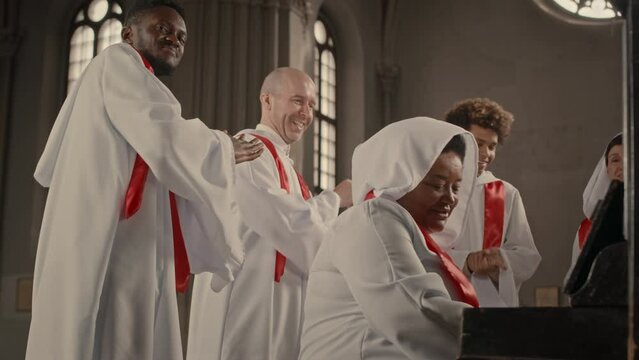 Multiethnic church choir wearing long white gowns making performance in Catholic Church. Mature Black woman playing piano while others singing and clapping their hands
