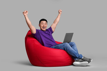 Emotional man with laptop sitting in beanbag chair against grey background