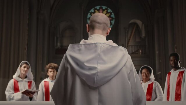 Back view of bald headed conductor speaking to multiethnic members of church choir while rehearsing for performance in old Catholic Church