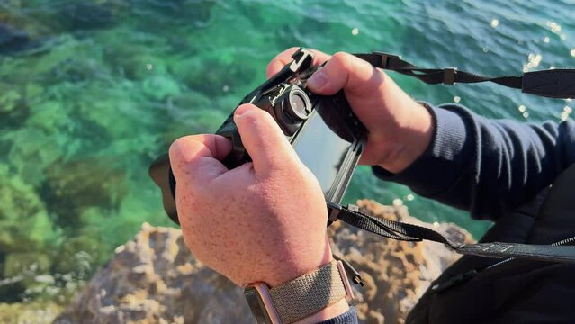 Hands of photographer hold camera on strap looks at screen photographs transparent azure sea water, rocky bottom from cliff, cliff. photojournalist makes report about nature. Tourist, Generative AI