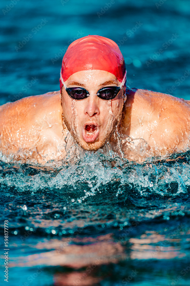 Wall mural Man Swimming. Male Swimmer athlete doing butterfly swim stroke. Male sport fitness man wearing swimming goggles and swim cap training hard in outdoor pool. Professional sports fitness model