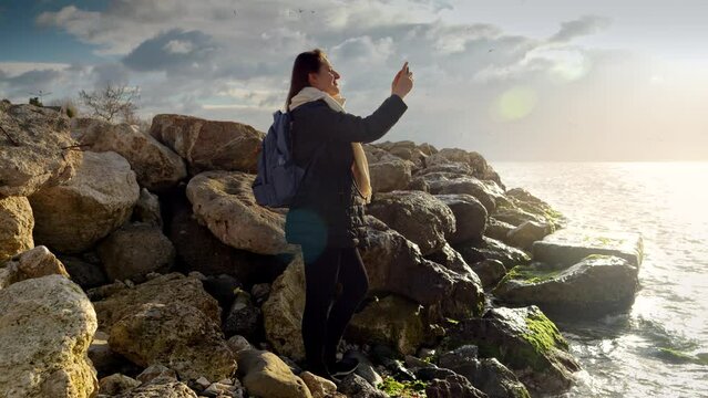 Beautiful young woman standing on the rocks at sea coast and making photographs of cold sea waves using smartphone. Concept of tourism, travel, journey, holiday and vacation at winter