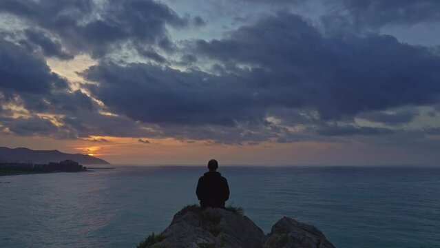 Timelapse captures lone man sitting on rock watching golden sunrise over the calm sea