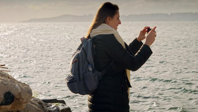 Young woman stands on the shoreline, capturing the winter ocean with her smartphone. Perfect for travel and vacation stock footage