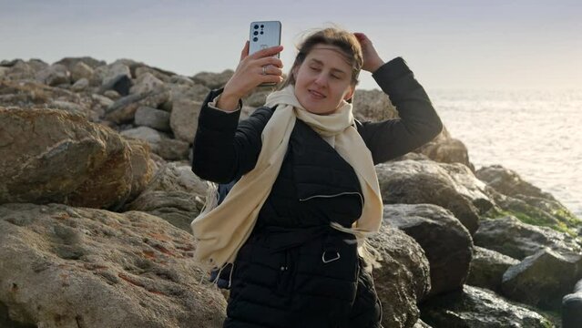 Young woman taking photos or herself at the winter sea using her smartphone while standing on the rocky shore. Perfect for travel and vacation footage
