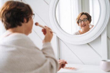 Smiling middle aged plus size woman doing makeup near mirror at home