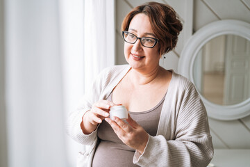 Smiling middle aged plus size woman with day cream near window at home