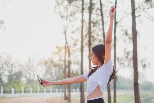 A beautiful female is using mobile phone to video calll or selfie while stretching in th early morning. Concept of city life or urban activity with technology. New normal life. Exercise and jogging