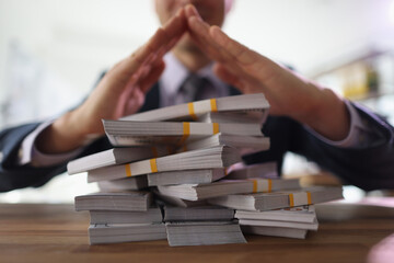 Businessman makes pyramid with hands above stacks of money