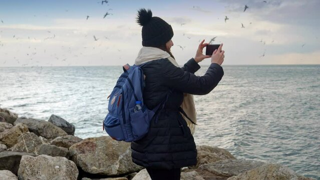 Woman in winter clothing, standing on the rocks by the sea, taking photos of the cold and stormy waves crashing against the shore with her smartphone. Travel, tourism, and adventure