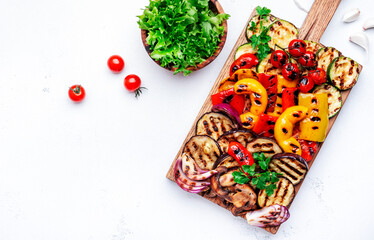 Grilled vegetables: red and yellow paprika, zucchini, eggplant, mushrooms, tomatoes and onions served on rustic wooden cutting board, white table background, top view