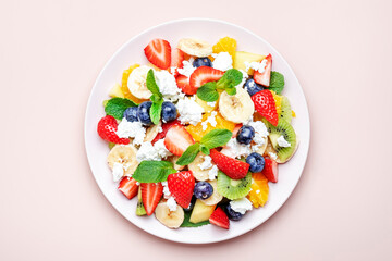 Sweet fruit berry salad with strawberries, blueberries, banana, soft cheese and mint leaves, pink plate on pink background, top view