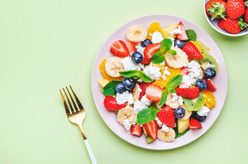 Summer fruit and berry salad with strawberries, blueberries, banana, soft cheese and mint leaves, green background, top view