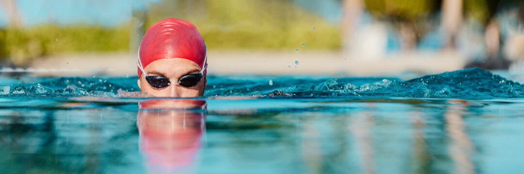 Swimming athlete man creative portrait wearing swim goggles and swim cap in swimming pool while doing breast stroke. Male swimmer swimming breaststroke in pool outside. Panoramic banner