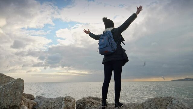A young woman with a backpack stands on a seaside cliff with her arms extended, enjoying the view of the cold ocean waves. This footage represents concepts of hiking, adventure, relaxation in nature, 