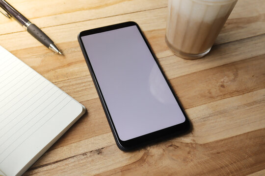 Blank screen mobile phone with empty notebook, pen, and a cup of coffee on wooden table during the day. Office work concept. Flat lay images