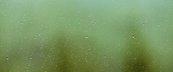 Atmospheric minimal backdrop with rain droplets on glass. Wet window with rainy drops and dirt spots closeup. Blurry minimalist background with greenery bokeh against dirty window glass with raindrops