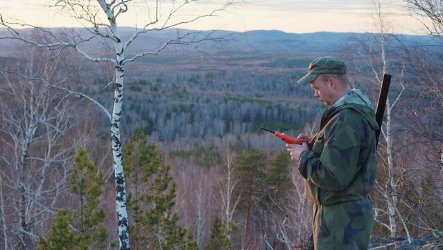 A hunter with a gun stands on the top of a mountain and looks at the location of his dog in a gps navigator. Hunting in the wild forest with a dog.