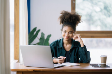 Happy black businesswoman using laptop to work and writing on notebook with pen in office with mobile next to laptop.