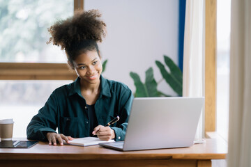 Happy black businesswoman using laptop to work and writing on notebook with pen in office with mobile next to laptop.