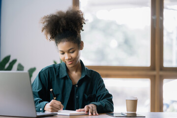 Happy black businesswoman using laptop to work and writing on notebook with pen in office with mobile next to laptop.